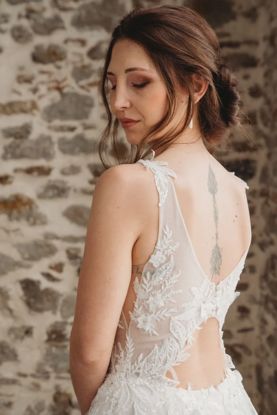 Mariée de dos, robe au dos en dentelle, devant un mur de pierre